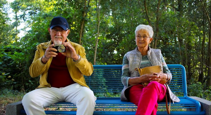 Dwoje starszych osób siedzących na ławce i robiących zdjęcia aparatem